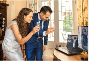 A joyful bride and groom share a celebratory champagne toast during a virtual wedding reception with loved ones on a laptop screen.